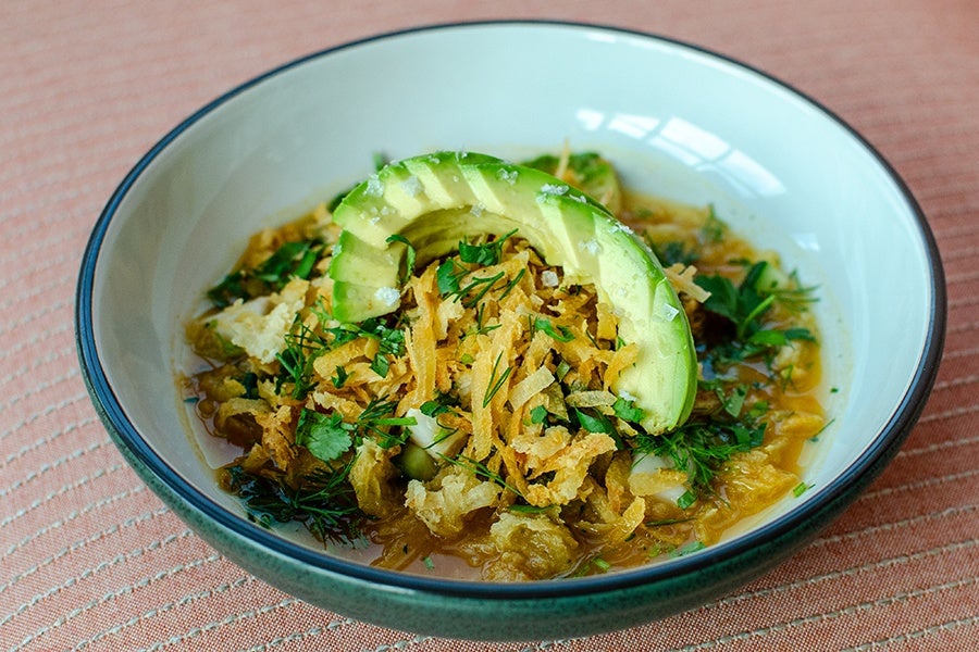 A bowl of ceviche garnished with a slice of avocado, crispy fried onions, and fresh chopped herbs, served in a white bowl with a dark rim on a textured pink surface.