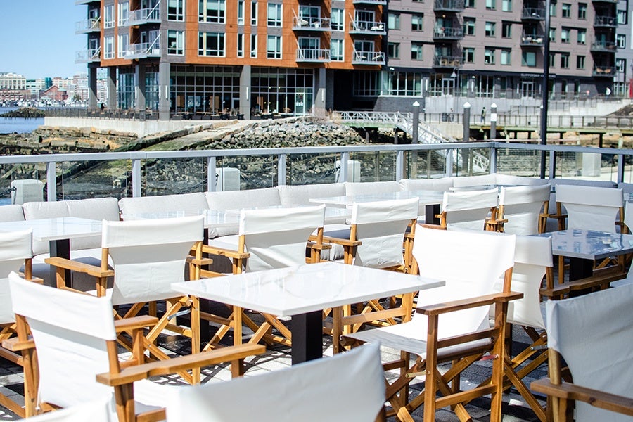 Outdoor seating area with white cushioned chairs and white tables on a patio overlooking a waterfront with modern buildings in the background.