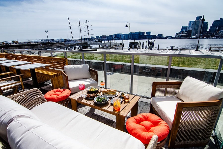 Outdoor seating area with cushioned wooden chairs and a sofa around a wooden coffee table set with plates of food and drinks, overlooking a waterfront with boats and city buildings in the background.