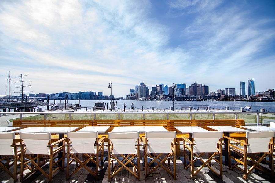 Outdoor seating area with wooden tables and white canvas chairs facing a waterfront, with a city skyline and partly cloudy sky in the background.