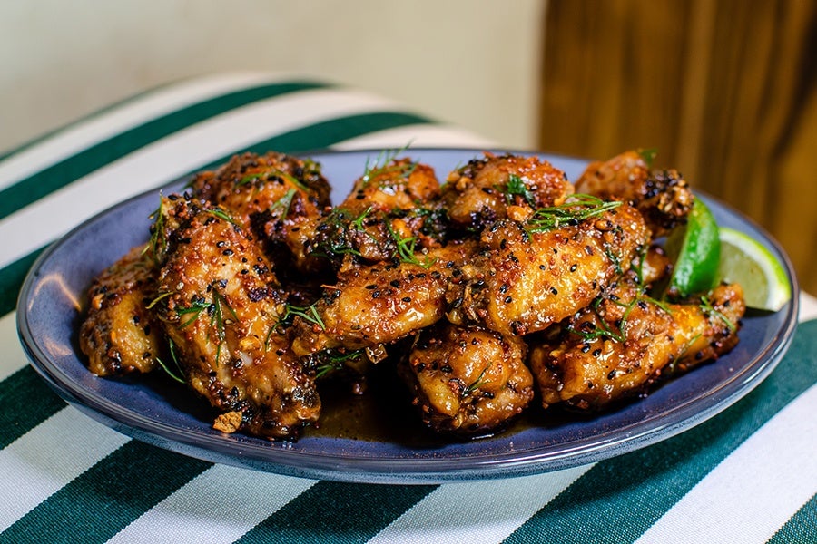 A blue plate filled with glazed chicken wings garnished with herbs and black sesame seeds, accompanied by lime wedges on the side. The plate is placed on a green and white striped cloth.