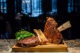 Two large, grilled bone-in steaks are presented on a wooden cutting board, accompanied by fresh green herbs. The steaks have a rich, charred crust and are positioned upright, showcasing the bones. The background is dark and blurred, highlighting the meat as the focal point.