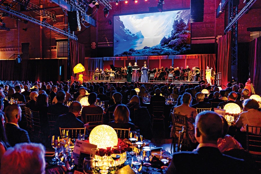 A formal indoor event with a large audience seated at round tables adorned with glowing spherical centerpieces. On stage, a group of musicians and singers perform in front of a large screen displaying a scenic image of a snowy mountain landscape. The room is dimly lit with warm lighting focused on the stage and tables.
