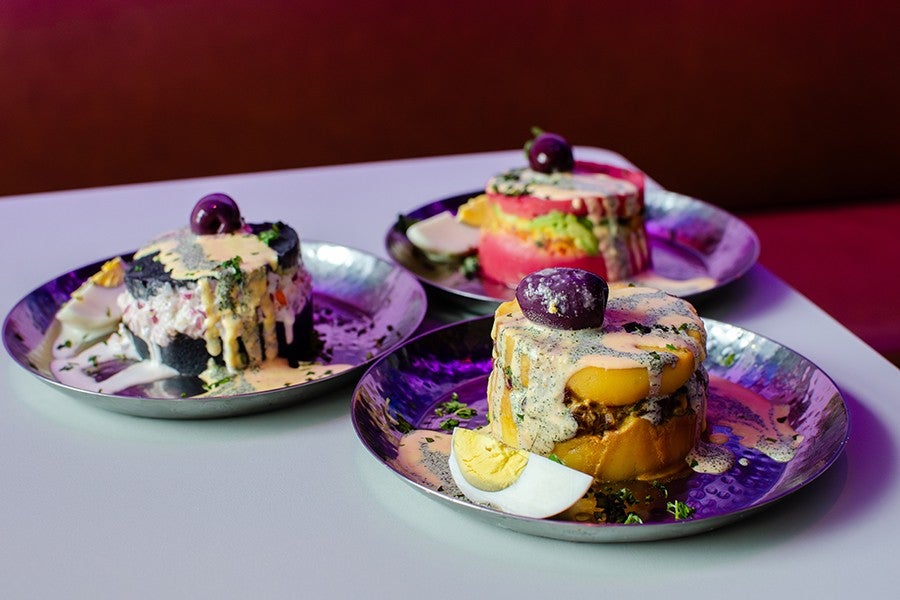 Three plated servings of causa, a layered Peruvian dish made with mashed potatoes and various fillings, each topped with a purple olive and drizzled with creamy sauce, accompanied by slices of hard-boiled egg on metal plates.