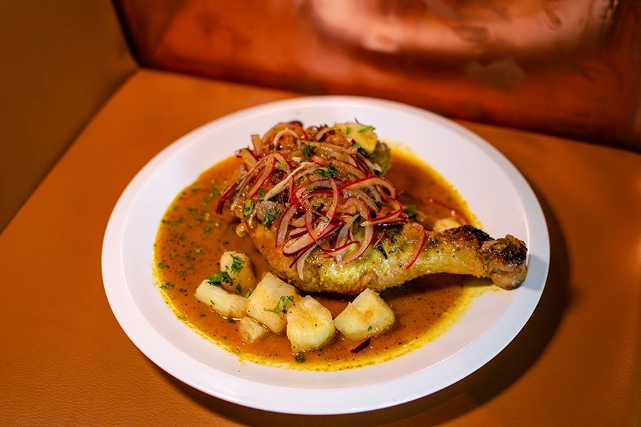 Roasted chicken leg topped with sautéed red onions and herbs, served with chunks of cooked potatoes in a brown sauce on a white plate.