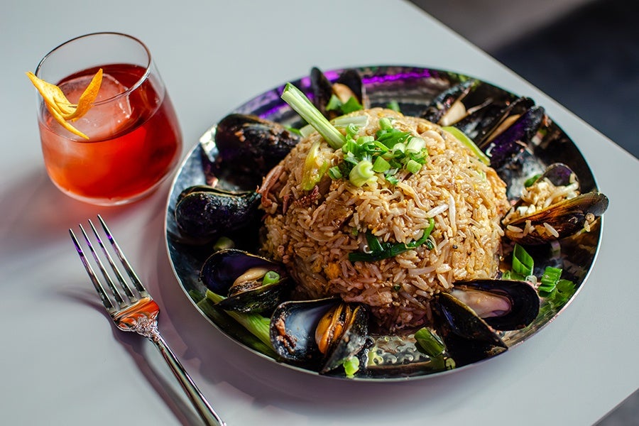A plate of fried rice garnished with chopped green onions and surrounded by mussels, served on a reflective metal plate. Next to the plate is a silver fork and a glass of reddish-orange cocktail with an orange peel garnish. The setting is on a light-colored table.