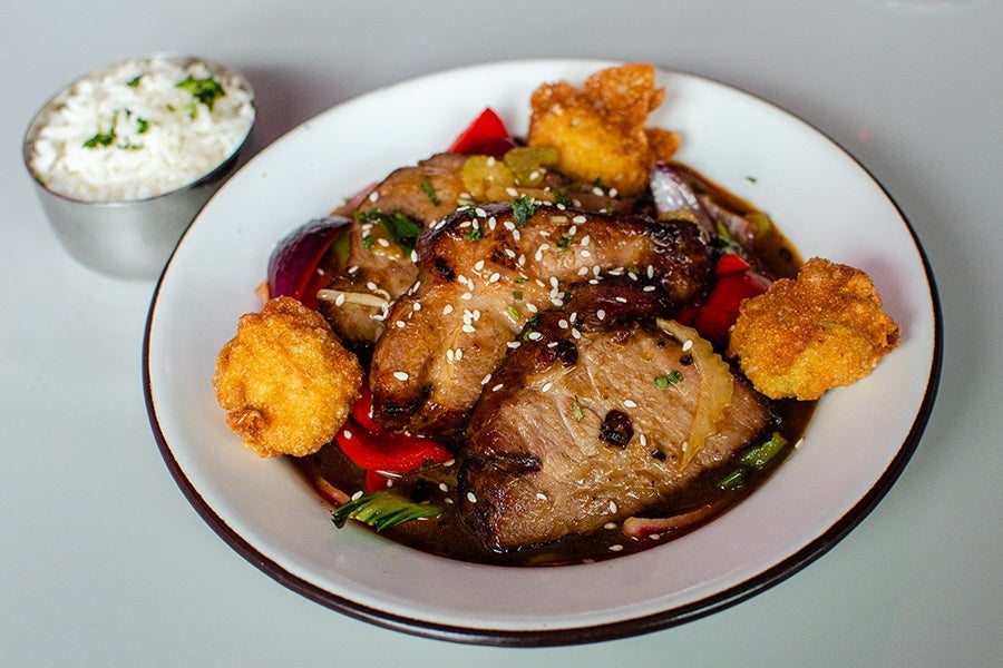 Grilled pork slices topped with sesame seeds, served on a bed of sautéed vegetables including red bell peppers and onions, accompanied by three pieces of fried battered cauliflower, with a side bowl of white rice garnished with chopped herbs.