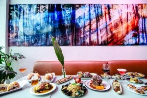A dining table set with a variety of dishes including sandwiches, seafood, and plated meals, accompanied by drinks such as beer, red wine, and cocktails. The table is in front of a brown leather bench with a tall green leaf in a glass vase as a centerpiece. Behind the bench is a large, colorful abstract artwork featuring vertical lines and a mix of blue, purple, and red hues. There are also green plants visible on the left side of the image.