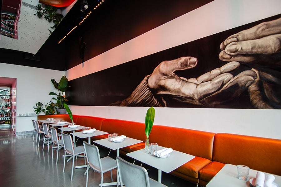 A modern restaurant interior with a long orange cushioned bench along the wall, paired with white tables and gray chairs. Each table is set with white napkins and glasses, and some have a single green leaf in a small vase. The wall above the bench features a large mural of two hands reaching toward each other on a black background. The floor is polished concrete, and there are plants near the far end of the seating area.