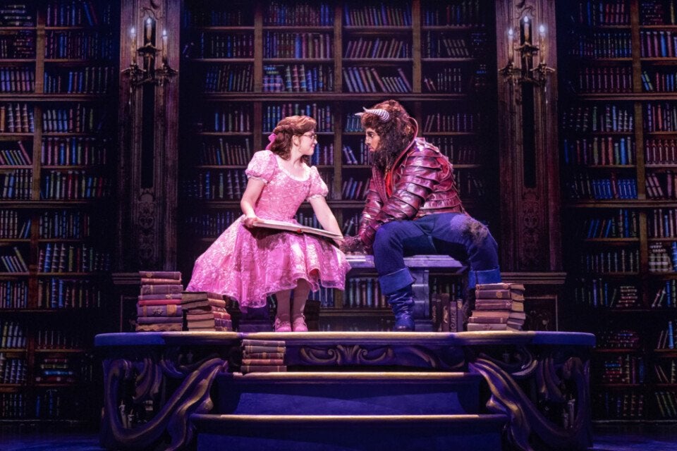 A theatrical scene featuring two characters sitting on a bench in front of a large bookshelf filled with books. One character is a woman wearing a pink, floral-patterned dress with puffed sleeves and pink shoes, holding an open book. The other character is dressed as a beast with horns, a mane, and a red armored jacket, wearing blue pants and boots. There are stacks of books on either side of the bench and on the steps in front. The setting is richly detailed with dark wood and warm lighting.
