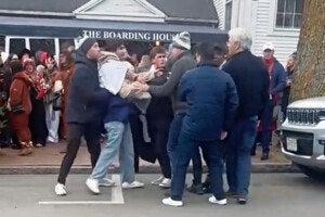 A group of men are engaged in a physical confrontation on a street in front of a building with a sign that reads "THE BOARDING HOUSE." Several people are watching the scene from the sidewalk, and a white vehicle is parked nearby. The men involved are wearing casual winter clothing, including jackets and hats.
