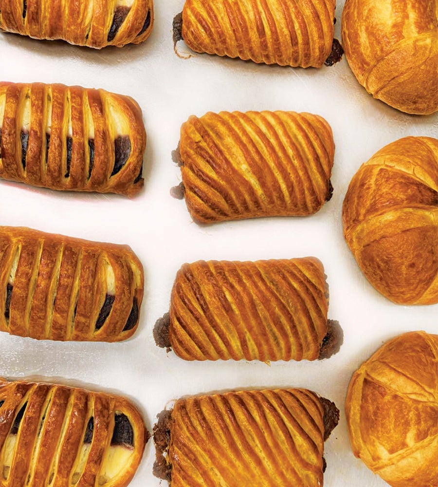 Golden-brown pastries with a flaky, layered crust, some with diagonal slits revealing a dark filling, arranged on a white surface. The pastries vary slightly in shape, with some rectangular and others round.