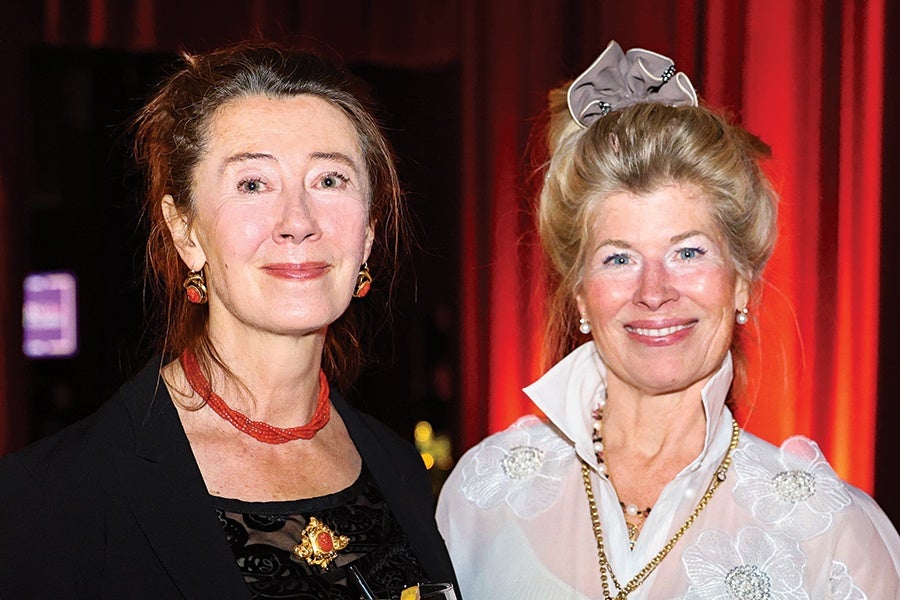 Two women are pictured against a dark background with red lighting. The woman on the left has dark hair pulled back and is wearing a black outfit with a red beaded necklace and matching earrings. The woman on the right has blonde hair styled up with a gray bow and is wearing a white blouse with floral embroidery and pearl earrings, along with a gold chain necklace. Both women are smiling.