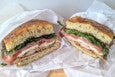 Two halves of a sandwich on focaccia bread, filled with chicken, fresh tomato slices, leafy greens, prosciutto, and mozzarella cheese, wrapped in white parchment paper.