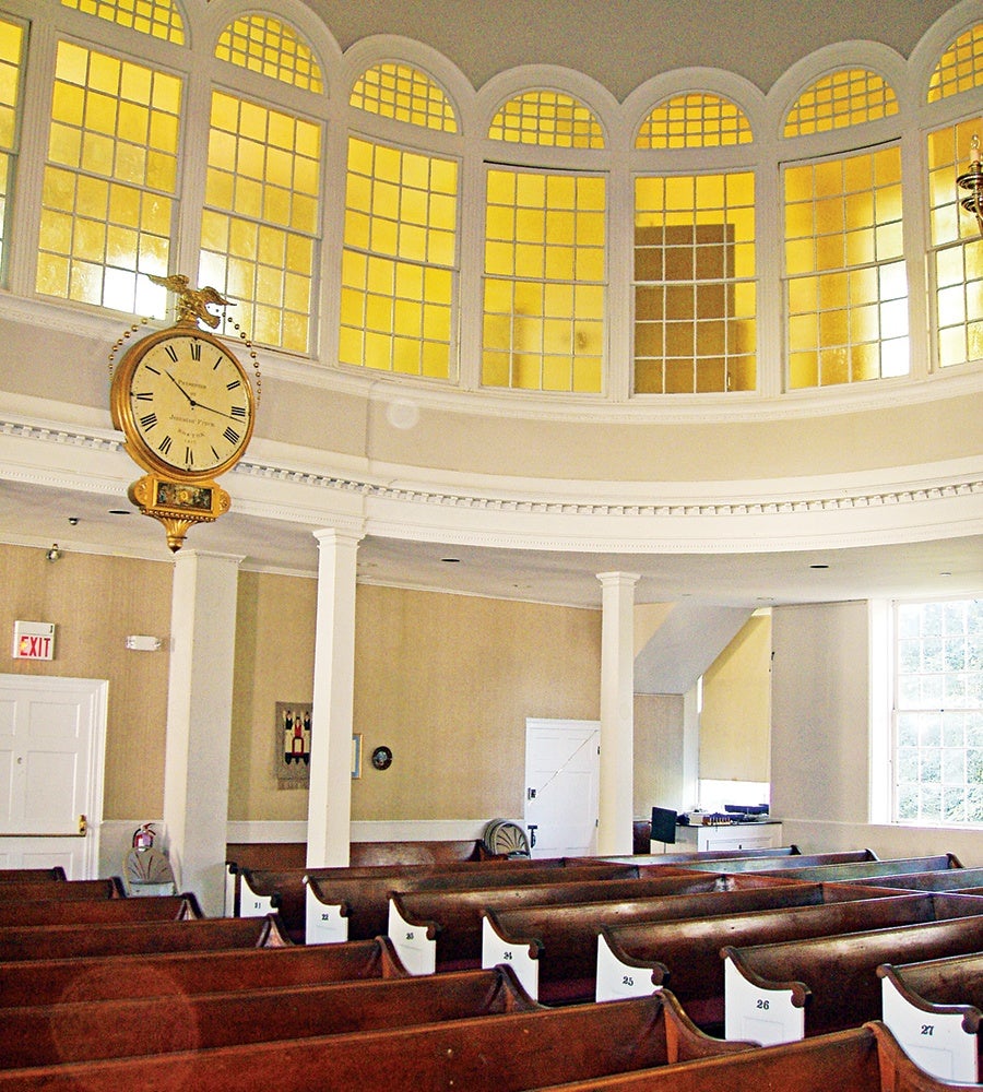 Interior of a historic church or meeting hall with wooden pews numbered 21 to 27, white columns, and a large round clock with Roman numerals hanging from the ceiling. The upper walls feature a series of tall, arched windows with yellow-tinted glass.