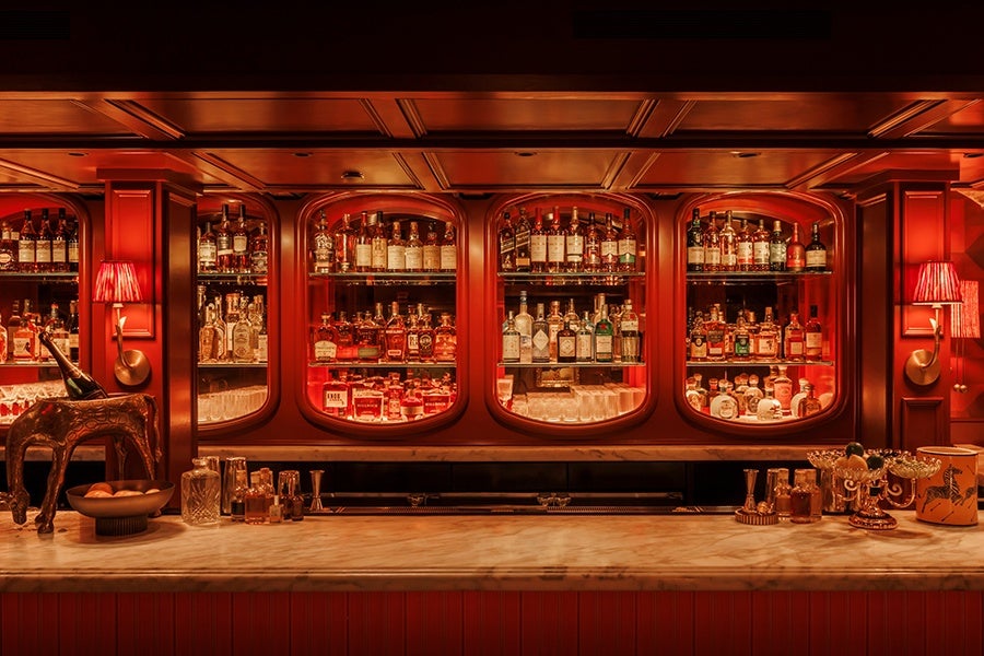 A warmly lit bar with a marble countertop and wooden cabinetry featuring four arched glass shelves filled with various liquor bottles. On the left side of the counter, there is a decorative horse sculpture holding a bottle, a bowl with oranges, and glassware. On the right side, there are ornate glassware and a decorative container. Two red wall lamps with pleated shades flank the shelving, enhancing the cozy, vintage ambiance.