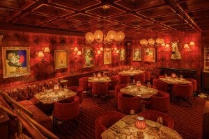A warmly lit, cozy restaurant interior with red patterned walls and carpet. The seating includes red upholstered chairs and banquettes around marble-topped tables set with glassware and napkins. The walls are decorated with framed abstract and figurative paintings, wall sconces with lampshades, and a zebra head sculpture. The ceiling features a geometric wooden design with hanging spherical paper lanterns.