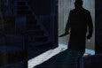 Silhouette of a person holding a large knife, standing in a dimly lit room with wooden panel walls, a staircase, and dark furniture. Light casts the shadow on the floor, creating a tense and ominous atmosphere.