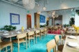 A modern dining area with light wooden tables and chairs featuring tan leather seats. The tables are set with white plates, napkins, and clear glasses, and each table has a small yellow lamp. The floor has a bright turquoise rug under the main table and a red rug reflected in a large wall mirror. The walls are white with colorful artwork, and there are several green plants around the room. Woven pendant lights hang from the ceiling, adding a cozy ambiance.