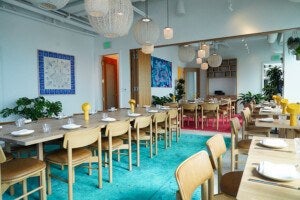A modern dining area with light wooden tables and chairs featuring tan leather seats. The tables are set with white plates, napkins, and clear glasses, and each table has a small yellow lamp. The floor has a bright turquoise rug under the main table and a red rug reflected in a large wall mirror. The walls are white with colorful artwork, and there are several green plants around the room. Woven pendant lights hang from the ceiling, adding a cozy ambiance.