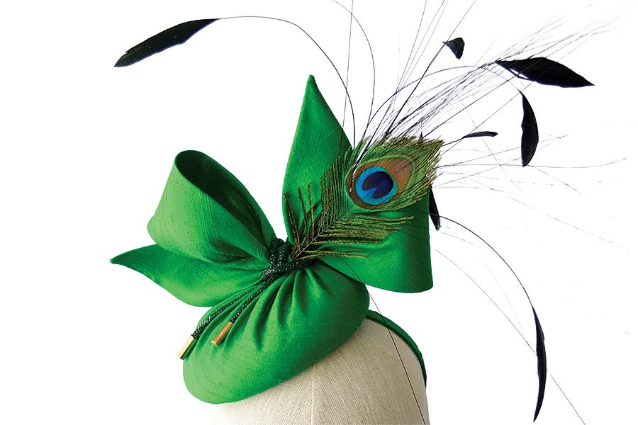 Green fascinator hat featuring large, sculpted fabric loops and a prominent peacock feather with additional thin black feathers extending outward. The hat is displayed on a beige mannequin head against a white background.