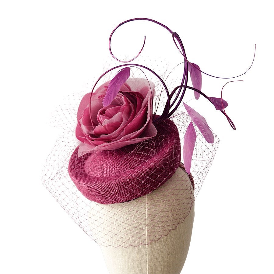 Small magenta fascinator hat featuring a large fabric rose, delicate netting veil, and decorative curled feathers.