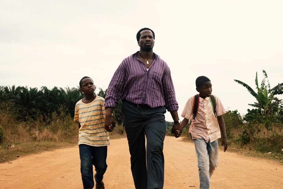 A man wearing a purple striped shirt and dark pants walks on a dirt road holding hands with two boys. The boy on the left wears a yellow and white striped t-shirt and dark jeans, while the boy on the right wears a light pink button-up shirt, light jeans, and a backpack. The background features greenery and an overcast sky.