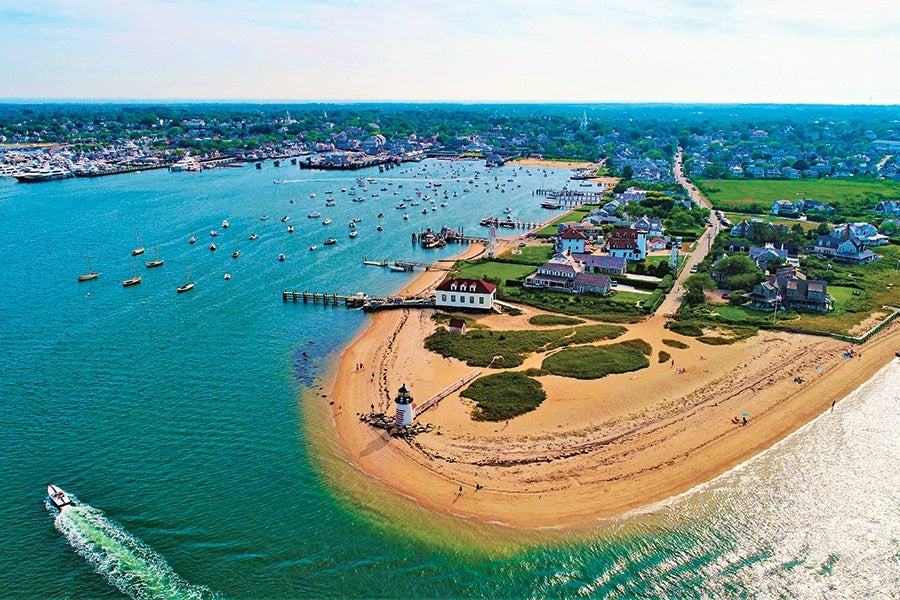 A coastal town with a sandy beach and a small lighthouse at the tip of a peninsula. Several houses and green patches are visible near the shore, with a road running through the area. Numerous boats are anchored in the calm blue water, and a boat is seen moving away, leaving a white wake behind. The town extends inland with more houses and greenery.