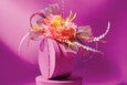 A vibrant floral headpiece featuring a large, multi-layered flower in shades of orange and yellow at the center. Surrounding the flower are delicate, translucent pink mesh loops and various colorful, thin, curly and zigzagged decorative elements in pink, yellow, blue, and white. The headpiece is displayed on a round, pink stand against a solid pink background.
