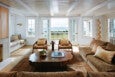 Living room with a large brown sectional sofa adorned with beige and brown pillows, two wooden armchairs with beige cushions, and a rustic wooden coffee table with books and a small plant. The room features white walls, large windows, and French doors that open to a balcony with a view of the ocean and greenery. A built-in bench with cushions is positioned under one window, and a modern wooden floor lamp stands near the sofa. The space is bright with natural light and has a cozy, coastal aesthetic.