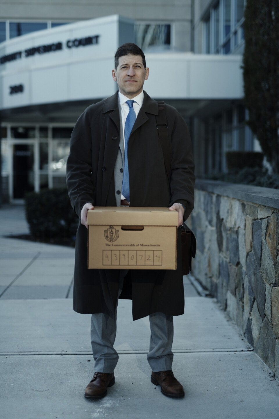 Man in a dark overcoat, gray suit, blue tie, and brown shoes standing outside a courthouse holding a cardboard box labeled "The Commonwealth of Massachusetts" with a number written on it.