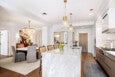Modern kitchen and dining area featuring a large marble island with a built-in sink and two pendant lights above. The dining area has a round table with six upholstered chairs, a crystal chandelier, and a sideboard with decorative items. The space has white walls, wooden flooring, and a wooden door at the back. Two rectangular mirrors hang on the wall near the island.