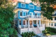 Three-story blue house with white trim and railings, featuring a front porch with stairs leading up to it, multiple windows, and surrounded by greenery and neighboring houses.