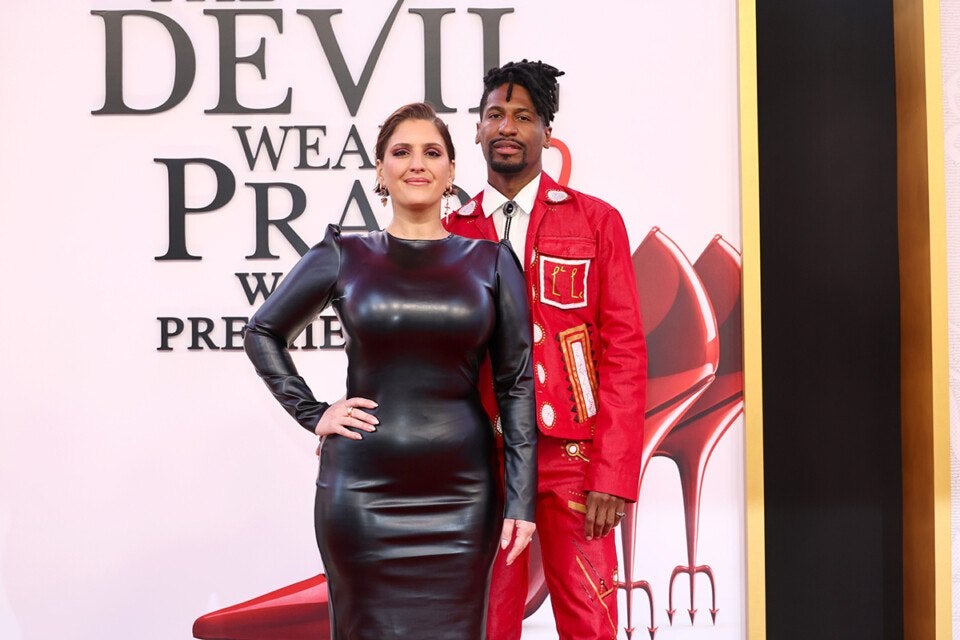 A woman in a fitted, long-sleeve black leather dress stands next to a man wearing a red suit adorned with white and gold patches. The man also wears a white shirt with a bolo tie. They are posing in front of a backdrop with large text and an image of red high-heeled shoes with devilish pitchfork heels.