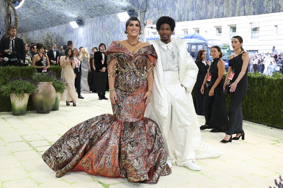 A woman and a man stand together on a light-colored tiled floor at an event. The woman wears an off-the-shoulder, floor-length gown with a voluminous skirt, featuring an intricate paisley pattern in shades of orange, brown, and gold, accessorized with gold jewelry. The man wears an all-white ensemble consisting of a long, puffy coat, a ruffled shirt, high-waisted pants, and white shoes. Behind them, several people dressed in formal black attire and other guests are visible, along with greenery and a blue bus outside.