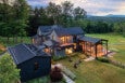 Modern multi-level house with dark exterior walls and metal roofs, featuring solar panels on the main roof. The house has large windows, some illuminated from inside, and a spacious glass-enclosed porch or sunroom. It is surrounded by a well-maintained lawn with outdoor seating around a fire pit, bordered by trees and open fields with distant hills under a partly cloudy sky.