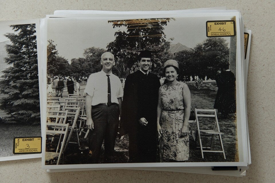 A black-and-white photo of three people standing outdoors on a grassy area with folding chairs in the background. The person in the center is wearing a graduation cap and gown, smiling. To the left is a man in a short-sleeve white shirt, dark tie, and dark pants, also smiling. To the right is a woman in a patterned sleeveless dress and a hat, smiling. Trees and other people are visible in the background. The photo has an "EXHIBIT 46" label in the top right corner.