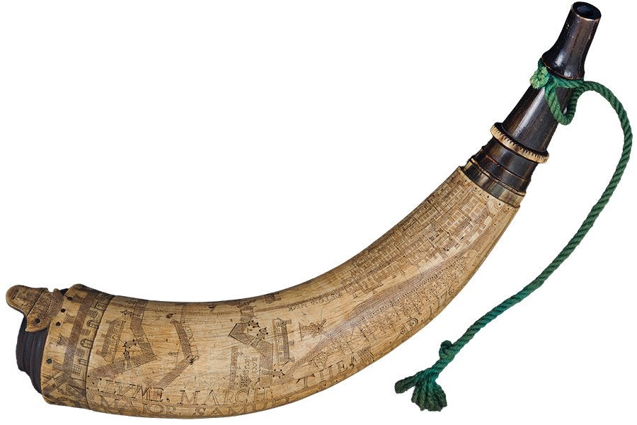 A curved powder horn made from a light-colored animal horn, featuring detailed engraved designs and text along its surface. The horn has a dark wooden stopper at the narrow end, secured with a green braided cord tied around it. The wider end also has a dark wooden base with a small protruding handle. The engravings include architectural elements and possibly text or names.