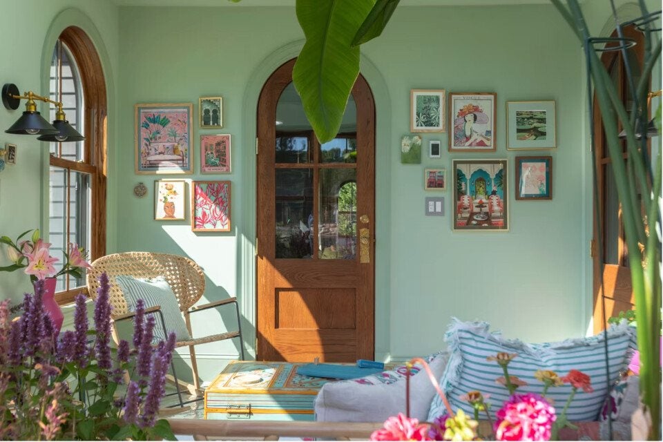 Sunlit room with mint green walls featuring a wooden arched door and windows. The walls are decorated with various framed artworks. A wicker chair with a striped cushion sits next to a colorful chest used as a coffee table. The foreground includes vibrant flowers and a striped pillow on a sofa.