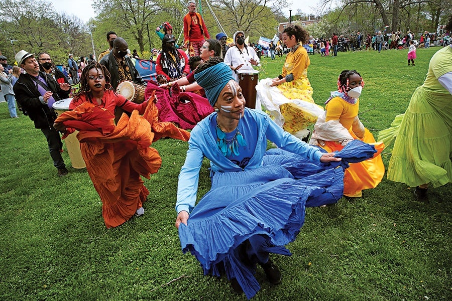 A group of people dancing outdoors on grass, wearing colorful flowing skirts and vibrant clothing. The central figure wears a blue outfit with a matching headwrap and face paint. Other dancers wear orange, yellow, and green skirts, with some also wearing face masks. In the background, musicians play drums, and more people are gathered, suggesting a lively cultural or festive event.