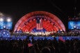 A large outdoor stage with a red, star-patterned backdrop and an American flag hanging in the center. The stage is filled with a choir and musicians dressed in black and white. Two large screens on either side display "The Herb Chambers" logo. The audience in the foreground is silhouetted, some holding small American flags. The scene is illuminated with stage lights against a dark night sky.