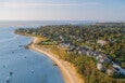 A coastal residential area with numerous houses surrounded by trees and greenery, bordered by a sandy beach and calm blue water with several small boats anchored nearby. The scene is captured under a clear sky with soft sunlight.