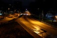 A dimly lit urban road at night with streetlights casting a yellow glow on the wet pavement. A single car is driving away in the distance. Trees line the right side of the road, and buildings are visible on the left. The road appears mostly empty and quiet.