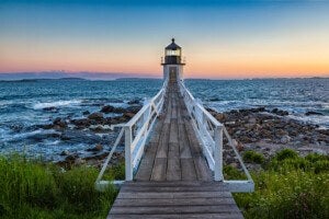 A wooden walkway with white railings leads to a small white lighthouse situated on rocky shorelines, with the ocean and a clear sky at sunset in the background. Green grass and wildflowers frame the foreground.