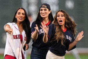 Three women wearing Boston Red Sox jerseys are engaged in an animated moment, with the woman on the left pointing forward, the woman in the middle smiling and gesturing with her hand, and the woman on the right appearing surprised or expressive with her mouth open and hands raised. The background is blurred, suggesting they are on a sports field.