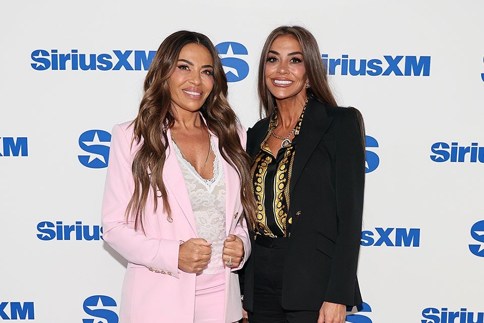 Two smiling women stand in front of a white backdrop with blue "SiriusXM" logos. The woman on the left wears a light pink suit with a white lace top underneath, and has long wavy brown hair. The woman on the right wears a black blazer over a black and gold patterned blouse, with long straight brown hair.
