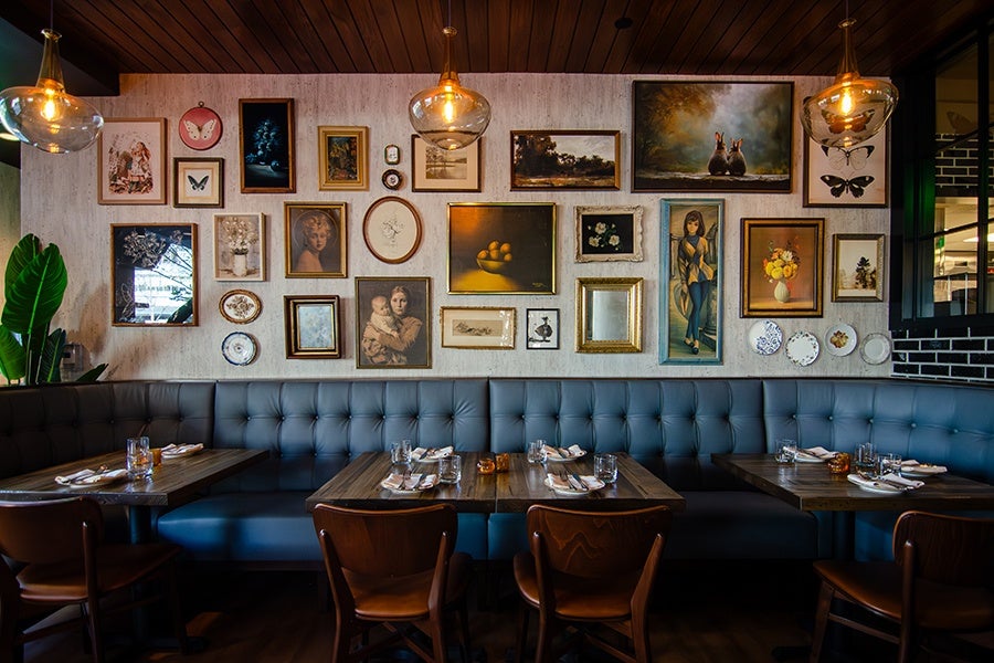 A cozy restaurant seating area with a long blue tufted bench against a wall adorned with a diverse collection of framed artwork and decorative plates. The tables are set with glasses, napkins, and cutlery, and wooden chairs are arranged around the tables. Three pendant lights with warm bulbs hang from a wooden ceiling, casting a soft glow over the space. Large green leaves peek out from the left side, adding a touch of greenery.