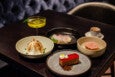 A dark wooden table with five items: a yellow cocktail in a coupe glass garnished with a green leaf and a small white flower, a toasted meringue dessert in a beige bowl, a rectangular layered chocolate and red jelly dessert on a gray plate with a small dollop of cream and a yellow decoration, a dish with a pink layered dessert in a beige bowl, and a small white cup of espresso on a saucer.