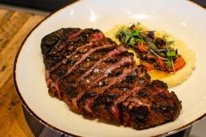 Grilled steak sliced and arranged on a white plate with a side of creamy polenta topped with roasted cherry tomatoes, herbs, and microgreens. The steak has visible grill marks and is seasoned with coarse salt.
