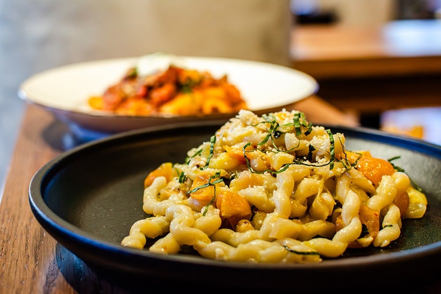 A black plate with twisted pasta mixed with yellow cherry tomatoes, grated cheese, and chopped fresh herbs, placed on a wooden table. In the background, there is a blurred white plate with another pasta dish.